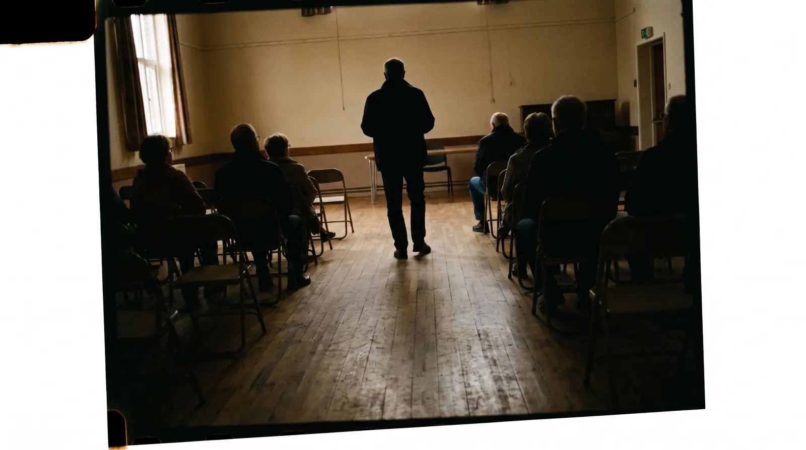 A person standing at the back of a dim room, watching a small gathering seated ahead