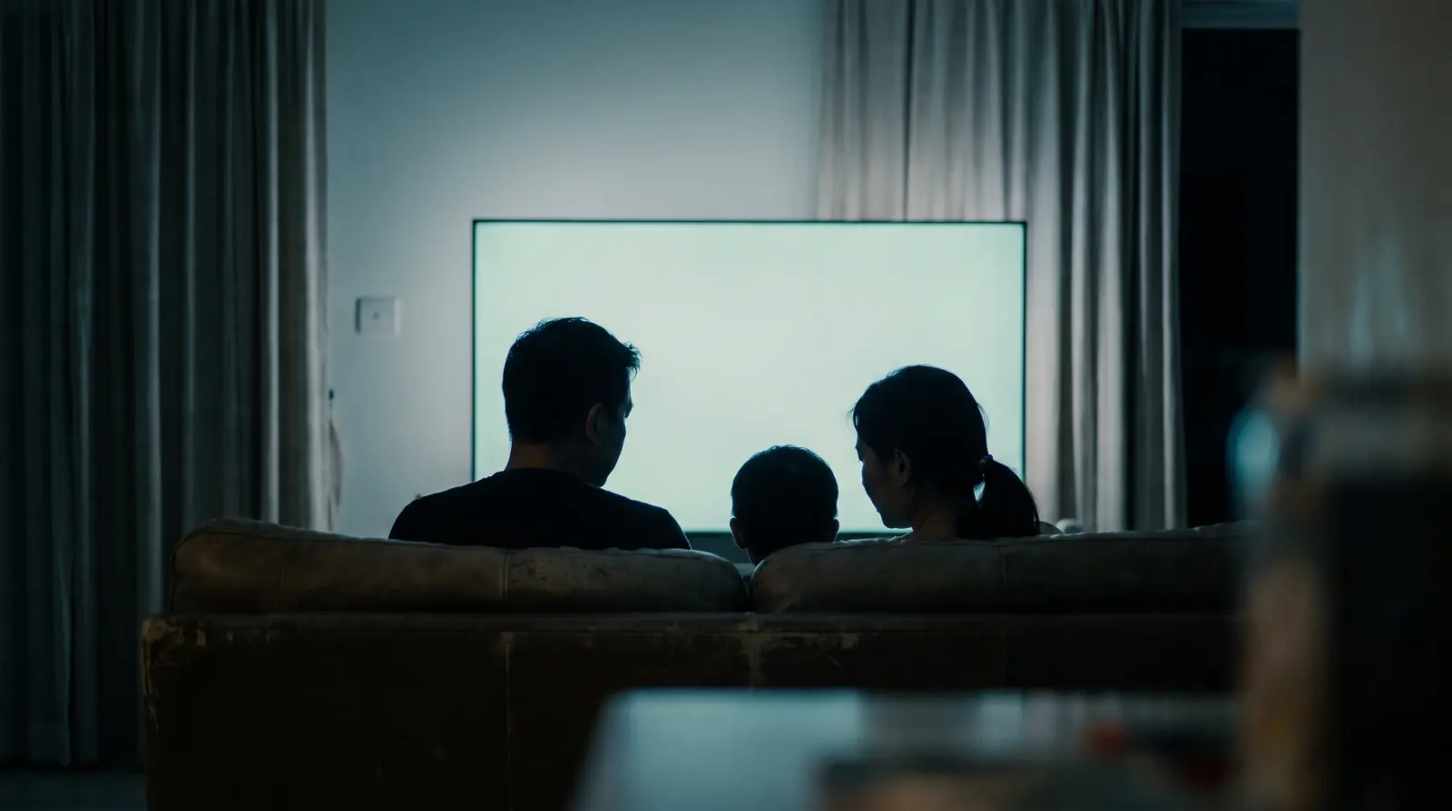 A family seated together in a dim room facing a softly lit screen, seen from behind