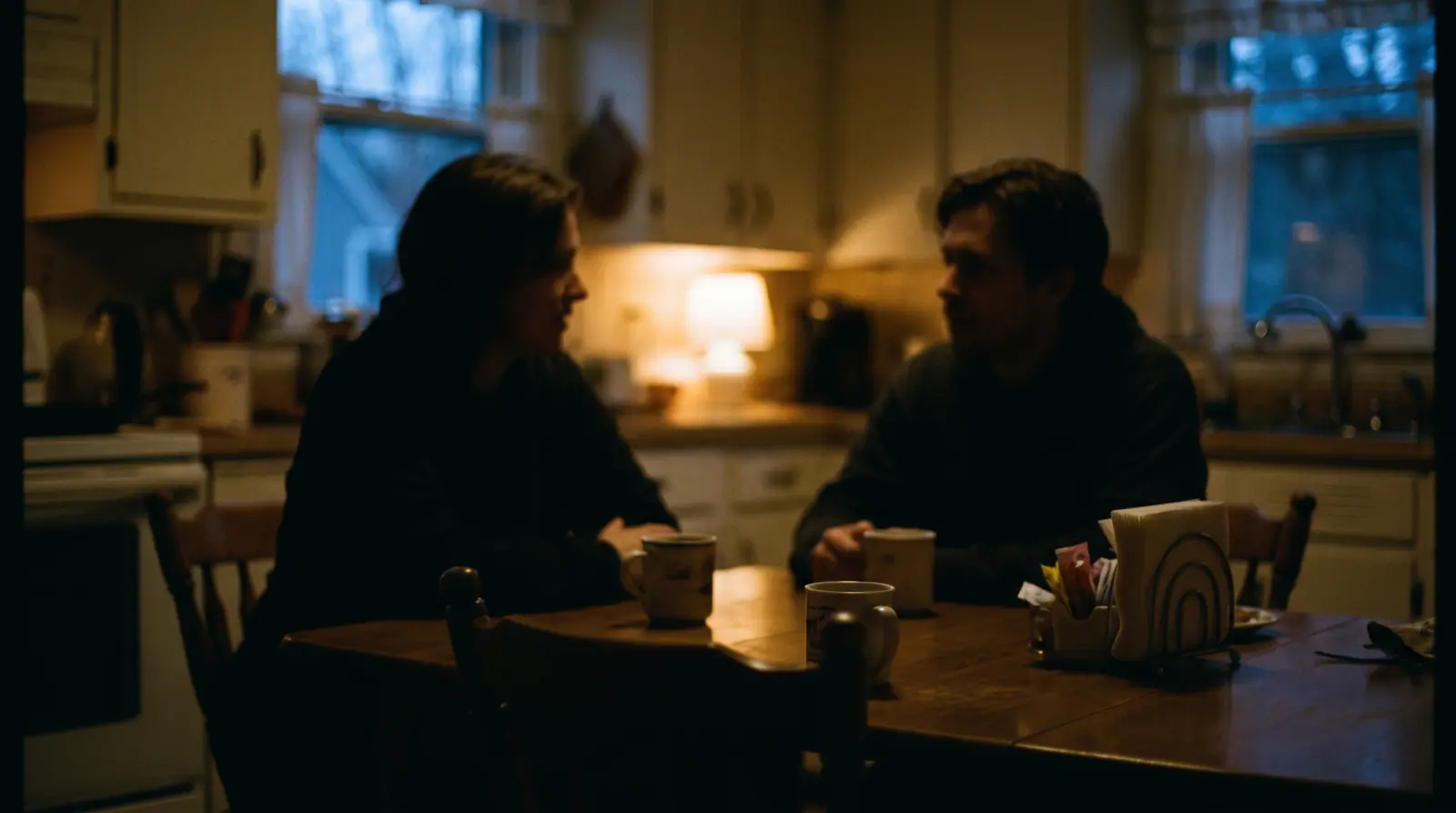 Two figures sitting across a kitchen table in evening light, talking