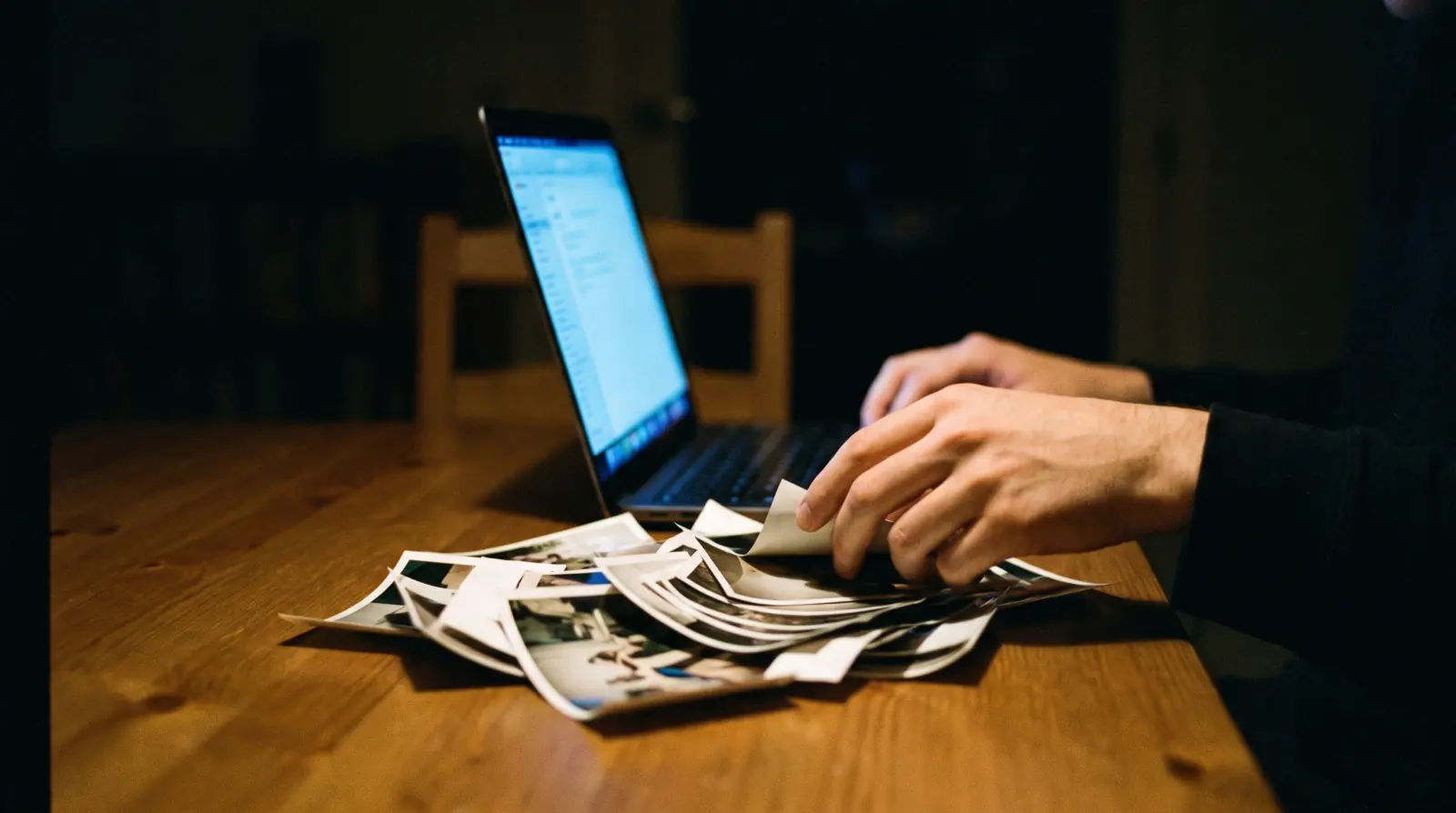 Hands working at a laptop late at night, scattered photographs lit by the screen